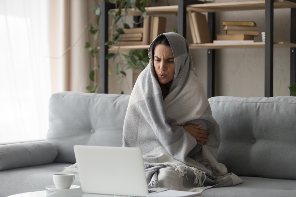 Woman sitting on the couch covered with the blanket shivering because heater is blowing cold air.
